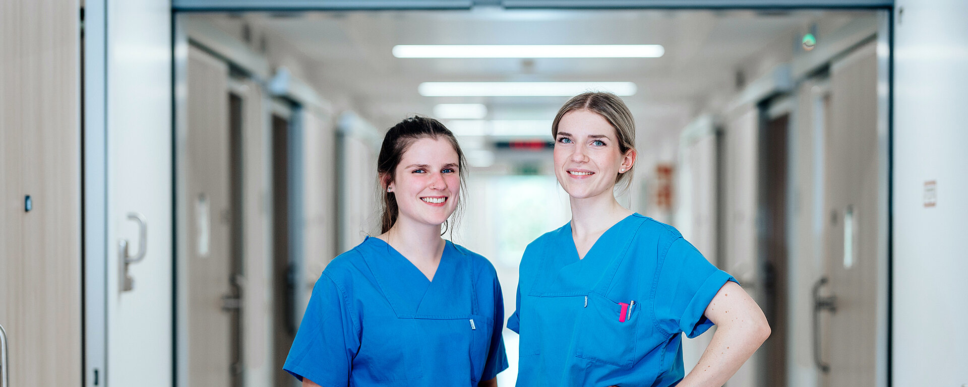 Zwei lächelnde Pflegefachkräfte, im Hintergrund der Krankenhausflur